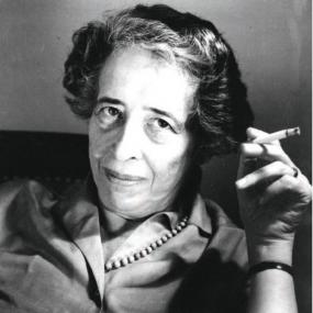 Black and white photograph of Hannah Arendt, leaning casually in a chair, with a raised cigarette in one hand. Her hair is short and she is wearing a button-up shirt and a beaded necklace.