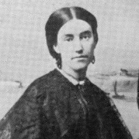 Black and white photograph of Mary Anne Barker. She is seated, eyes cast down, apparently in mourning since her dress and shawl are black. Her dark, gleaming hair is centre-parted and pinned back.