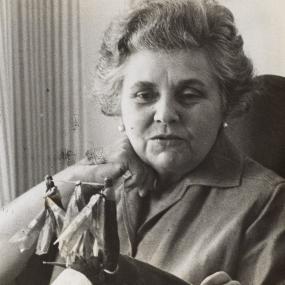 Black-and-white photograph of Elizabeth Bishop in 1964. Her hair is short and wavy; her chin rests on one hand. She wears pearl ear studs and a shirt with notched collar. In front of her stands an object which might be a group of miniature national flags.