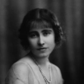 Black and white, head-and-shoulders photograph of Lilian Bowes Lyon, seen from the side with her face turned to face the viewer. She is wearing a light, lacy dress with short sleeves, and two strands of pearls. Her dark hair is parted in the middle, with a fringe.