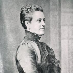 Black and white photograph of Anna Brassey, shown from the side. She is wearing a dress with a large bustle and ruffles at the edge of the sleeves, and she has a ruffled scarf around her neck. Her hair is pinned at the back of her head.