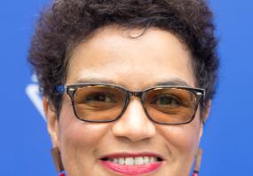 Photo of Jackie Kay taken during the annual Edinburgh International Book Festival. She is wearing brown tinted glasses and a dress with small
            polka-squares that matches her earrings. she has short black hair.