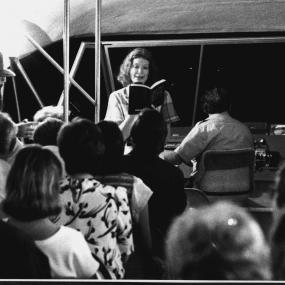 Black-and-white photograph of Fleur Adcock, seated with a microphone, reading her poetry to an audience aboard a covered boat during the
            Adelaide Festival of Arts, 5 March 1986.