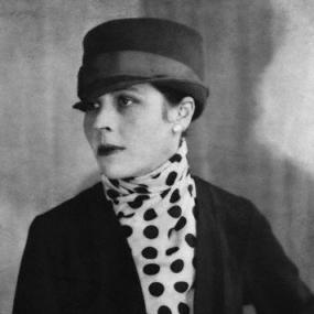 Black and white photograph of Djuna Barnes, shown from the waist up, with her head turned slightly and one hand on her hip. She is wearing a polka-dotted blouse under a black jacket, large pearl earrings, dark lipstick, and a black hat with a ribbon around it and a narrow brim.