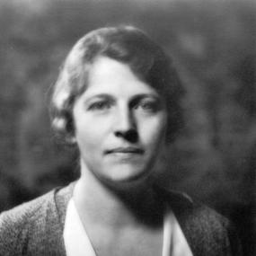 Black and white photograph of Pearl S. Buck, shown from the shoulders up. She is wearing a light dress and a dark cardigan, with her wavy hair pulled loosely back.