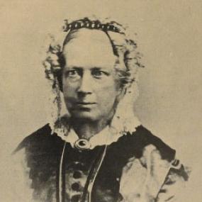Sepia photo of Mary Carpenter. She wears her grey hair in curls, under a head-band and cap with ear flaps. She has a dark, buttoned bodice with paler sleeves and white collar with a pendant in the centre.