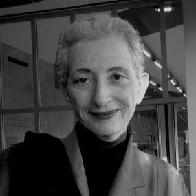Black and white photograph of Hélène Cixous by Claude Truong-Ngoc, September 2011. She is wearing a light jacket over a high necked black sweater, with a piece of black clothing swung over her shoulder. She has short, cropped grey hair and a slight, enigmatic smile.