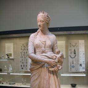 Marble statue of Anne Damer as the Muse of Sculpture by Giuseppe Ceracchi, 1778. She stands on a square plinth engraved with her name ("The Honourable Anne Seymour Damer") wearing classical Greek garb, with a flower wreath on her tightly-curled hair and a girdle below her breasts. She holds a statuette of a river god, with urn. British Museum.