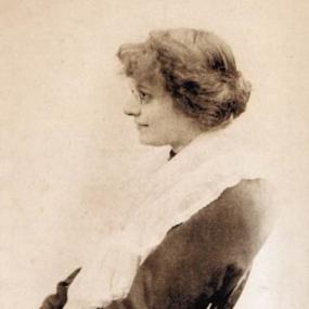 Black-and-white photograph of Eleanor Farjeon sitting on a chair looking left. She is wearing glasses and a dark dress with a white shawl draped over her neck. Her hair is short and wavy, and she is smiling slightly.