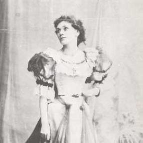 Black and white photo of Florence Farr standing with her left hand on her waist, looking off to the left. Her dress has a wide, long skirt, and large lacy ruffles at collar and cuffs. She seems to be standing in front of a curtain.