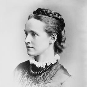 Head-and-shoulders photograph of Millicent Garrett Fawcett. She is looking to the left with her hair braided and coiled around her head, wearing a frilled collar and a necklace of large beads.