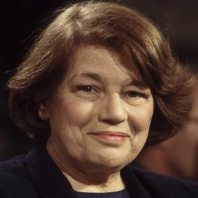 Colour portrait photo of Mavis Gallant in Paris, July 1984. She is smiling and looking directly at the camera. She wears a navy blazer with a
            brown stone brooch at the side and a small microphone at her collar. 