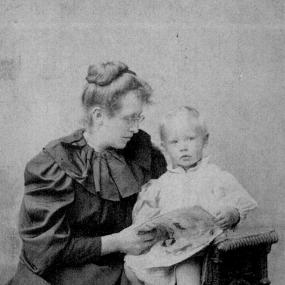Black and white photograph of Constance Garnett in the mid 1890s, sitting beside her young son David. She is turned towards him and together they hold a picture or a picture book.  She wears a long dark dress with dark gathered collar and small spectacles, and her hair is neatly pinned in a bun on top of her head.