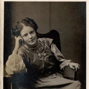 Sepia photograph of a young Mary Gawthorpe leaning sideways in an armchair with her head propped on one hand. She wears a blouse with full sleeves and a waistcoat over it. Her short, curly hair is centre-parted and brushed back. An inscription in capitals reads "Miss Mary Gawthorpe. National Women's Social and Political Union, 4 Clements Inn London W. C."