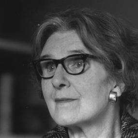 Black-and-white photo of Stella Gibbons, 28 November 1977. She is posing in front of bookshelves, looking off into the distance from behind
            tortoise-shell glasses. 