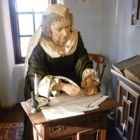 Colour photograph of a mannequin representing Anne Halkett, sitting at a desk writing, dressed in black with white cap, collar, and cuffs. The picture was taken at the heritage centre (since 2015 struggling for financial support) in Abbot House, Dunfermline, the oldest building in the town where Halkett spent her last three decades.