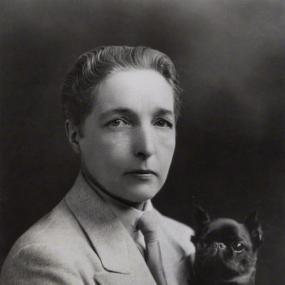 Black and white photograph of Radclyffe Hall, shown from the waist up. She is wearing a man's suit and tie, and her short hair is slicked back. In her left hand she holds a small black dog.