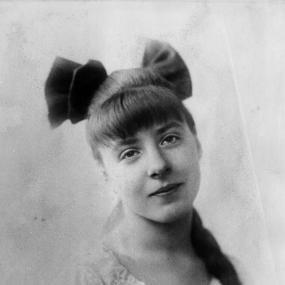 Black-and-white photo of Irene Handl, circa 1918. She has her hair in a braid with a large bow at the top and wears a blouse with a ruffled
            collar. The edges of her image blur into the white of the background.