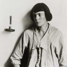 Black-and-white photo of H.D. posing, looking off-camera and leaning against a wall with her hands in her pockets. Her hair is styled in a
            page-boy cut. 