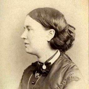 Sepia photograph of Jean Ingelow, shown in profile from the shoulders up. Her hair is in a bun at the nape of her neck; she wears a dress with buttons up the front and a high collar with a bow around it and necklaces.