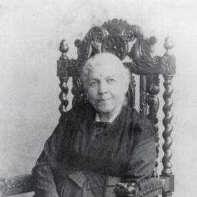 Black and white photograph of Harriet Jacobs, seated in an ornately carved, high-backed chair, wearing a long dark dress with long sleeves and a multi-layered dark bodice. Her white hair is frizzy and pulled back.