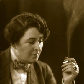 Black-and-white photo of Naomi Jacob, circa 1930. She wears a suit and bow tie, with her hair short and finger waved. She gazes at a lit
            cigarette in her left hand that is billowing smoke. 