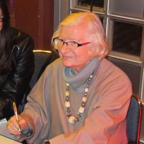 Photograph of P. D. James in Cologne, 2013. She sits with pen in hand, ready to sign a book. She is wearing a loose-fitting grey turtleneck, with a chunky necklace and a wristwatch, and glasses. Her grey hair is chin-length.