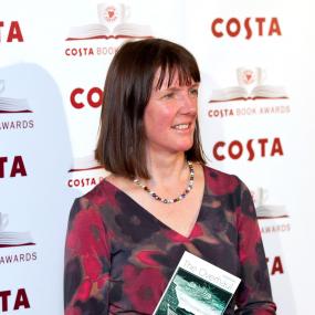 Colour photo of Kathleen Jamie holding a copy of her book The Overhaul at the Costa Book Awards in London, 29 January 2013. Standing and shown
            in profile, she wears a brown-, red-, and grey-patterned dress. 