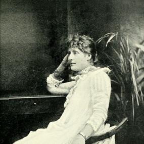 Black and white photograph of Harriett Jay, seated in a chair and leaning her elbow on a desk, with a potted plant behind. She is wearing a long, simple, white dress with lace trim at the sleeves and throat, and a silver bracelet and ring. Her light hair is pulled back, with short curls in front.