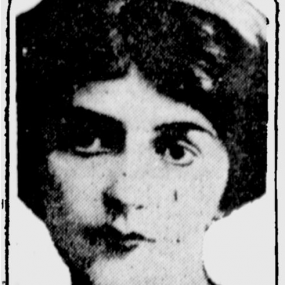 Black and white photograph of F. Tennyson Jesse, shown from the shoulders up, in a shape with symmetrically ornamental corners. The picture, with strong light and dark contrast, shows her wearing a small hat or headband on short, dark, bushy hair parted in the middle, and a white blouse with black bow.