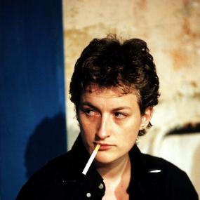Photograph of Sarah Kane. She is wearing a black button up and has short hair. She is holding an unlt cigarette in her mouth. 