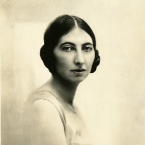 Black and white photograph of Margaret Kennedy. She is seated, with her body turned away but her head turned back to face the viewer. Her dark hair is coiled over her ears, and she wears a light dress with lace.