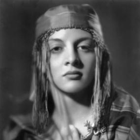 Black-and-white photo of Marghanita Laski. She is wearing a tassled cap and is draped in striped fabric. Therer is a small gecko brooch on her
            left that sits between her fingers. 