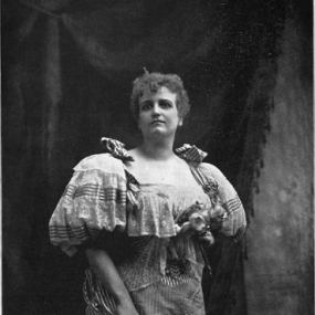Black and white, three-quarter-length photograph of Mary Lavin, standing with drapery behind her, seen slightly from below. Her hair is short and rough; she wears a short-sleeved, striped dress with bows on the shoulders and fabric roses over the bust.