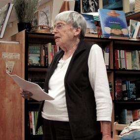 Colour photo of Ursula K. Le Guin, standing in a bookshop, apparently reading from papers in her hand, resting the other hand on a table. She has short grey hair and wire-rimmed glasses, and is wearing black pants, a white shirt, and a black vest.