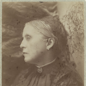 Faded black and white studio photograph of Katharine S. Macquoid by H. I. Mendelsohn of London. She is seen from the waist up against a backdrop of vegetation, turning with her face in profile. She wears a dark dress with a high collar closed with a brooch. Her smooth grey hair is pulled back and part covered by a black cap; she has a pearl stud in each ear. Her signature is written across the bottom of the picture.