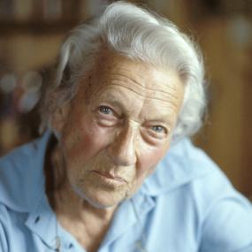 Portrait of Ella K. Maillart. She is wearing a light blue polo shirt. Behind her is a stack of books, blurred in the
            background.