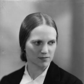 Black and white photograph of Ethel Mannin, shown from the shoulders up. She is wearing a dark suit over a light collared shirt, and her smooth hair is pulled back into a bun at the nape of her neck.