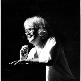 Black-and-white photo of Jan Morris speaking at a Herald literary luncheon in Sydney, Australia, 7 April 1992. Leaning on the podium, she is
            smiling at the audience.