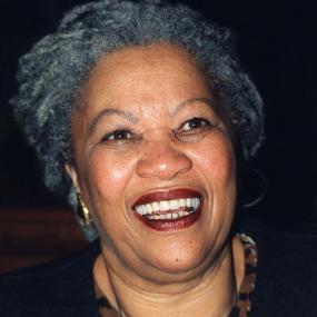 Head-shot in colour of Toni Morrison with a big smile. She is wearing red lipstick, gold earrings, and a black jacket over a leopard-spotted shirt. Her hair is short and grey.