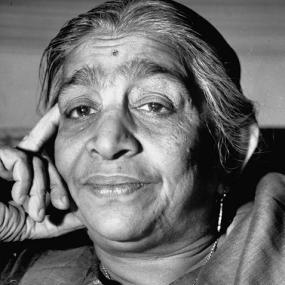 Black and white photo of Sarojini Naidu, head and neck only. She looks quizzically at the camera, her face resting on one hand, with slightly raised eyebrows emphasizing the wrinkles on her forehead. Her dark grey hair is brushed back, and she wears dangly earrings.