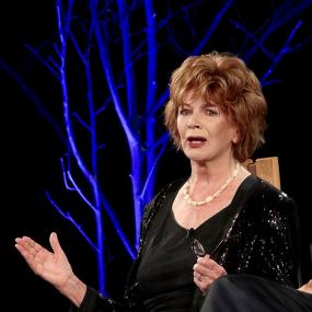 Colour photograph of Edna O&apos;Brien speaking at the Hay-on-Wye Festival in 2016.  She is seated on a wooden chair against a black backdrop with projected blue glowing trees. On a table in front of her are several books (including her recent "The Little Red Chairs"), a jug of water, drinking glasses, and a cloth of some kind. Her auburn hair is short and perkily styled; she wears berry-pink lipstick, a white chunky necklace, a black shirt and black sparkly cardigan, and holds her black-rimmed reading glas