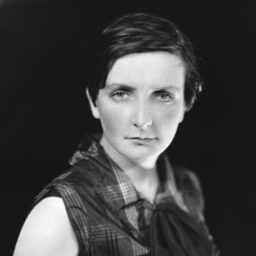 Black-and-white photo of Kate O'brien, 1926. Standing and looking intently at the camera, she is wearing a sleeveless plaid blouse with a tie
            at the neck and has her hair cropped short.
