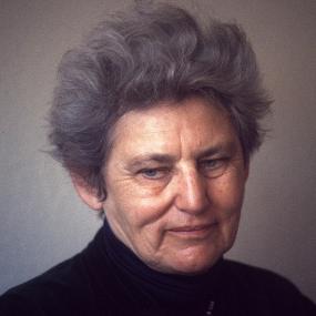 Photograph of Tillie Olsen, shown from the shoulders up, wearing a black turtleneck and a necklace of small red, blue, and white beads. Her hair is short, grey, and sticking straight up from her head.