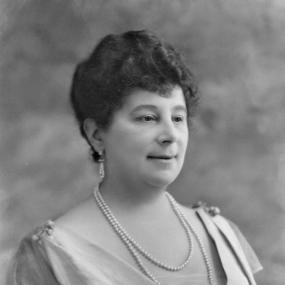 Black and white photograph of Baroness Emmuska Orczy, shown from the shoulders up, wearing a V-necked dress pinned with a brooch, with a gauzy fichu in the V and a larger brooch pinned in the middle. She is wearing pearl earrings and two strands of pearl necklace; her hair is dark, short, and curly.