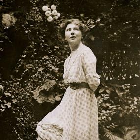 Black and white photograph of young Christabel Pankhurst, standing in a garden with roses and other flowering plants behind her. She has her hands behind her back and one knee bent under a long flowing dress with draped skirt over pleated under-skirt, and a thick dark waistband. She has jaw-length wavy hair and her face is turned up as if to look at the sky.
