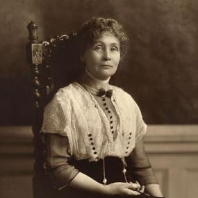 Sepia photograph of Emmeline Pankhurst, seated in a carved high-backed chair. She is wearing a white lacy button-up shirt with a high neck and bow at the collar, tucked into a black skirt with a high waist. She is wearing a long necklace, has short curly hair, and holds a pen in the hand which rests on her lap. Her expression is challenging.