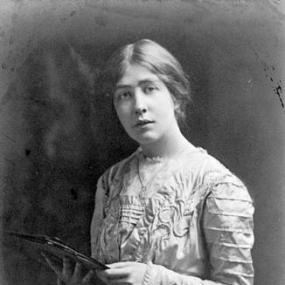 Black and white, half-length photograph of Sylvia Pankhurst, standing looking into the camera, holding a thin book. Her hair is centre-parted and loosely pulled back; she wear a long skirt and a top with long sleeves and embossed detail.
