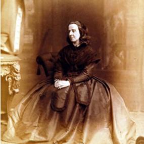 Sepia toned photograph of Bessie Rayner Parkes, seated on an upholstered chair, with an ornate table next to her. Her hands are clasped in her lap, she is wearing a dark dress with a voluminous skirt and long sleeves. She has dark, wavy, shoulder-length hair.