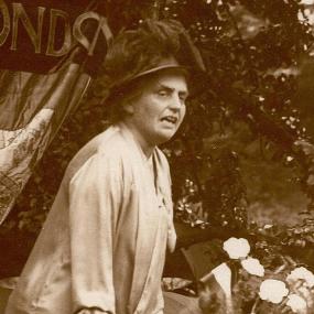 Sepia photograph of Eleanor Rathbone campaigning outdoors. She leans forward as she speaks, her hands on a table bearing a vase of white flowers, There are trees and a banner behind her. She is wearing, apparently, a loose-fitting white summer suit, and a black hat, elaborate but compact.